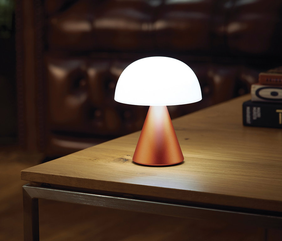 Eine orangefarbene LEXON LED-Akkutischleuchte »Mina M« steht auf einem Holztisch.