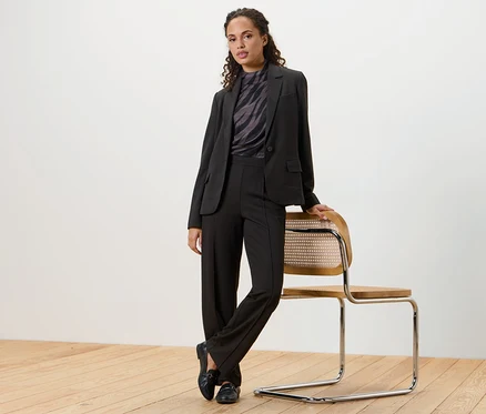 Eine Frau posiert in einem schwarzen Jerseyblazer und einer Hose und lehnt an einem Stuhl.