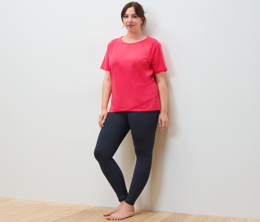 Eine Frau steht an einer Wand, sie trägt ein Sportshirt und eine Sporttight, navy.