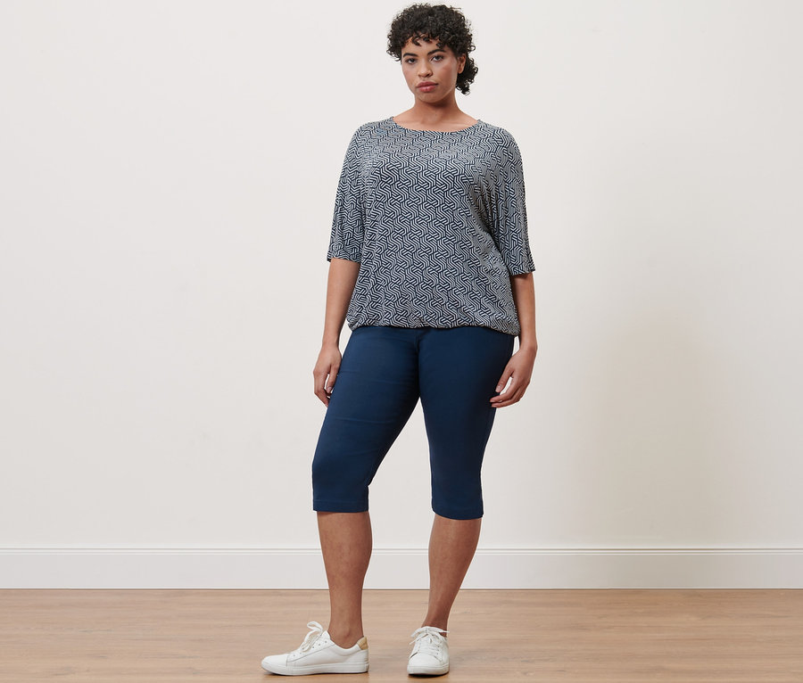 Frau steht und trägt ein blaues bedrucktes Blusenshirt und eine navyfarbene Bengalin-Hose in 3/4-Länge.