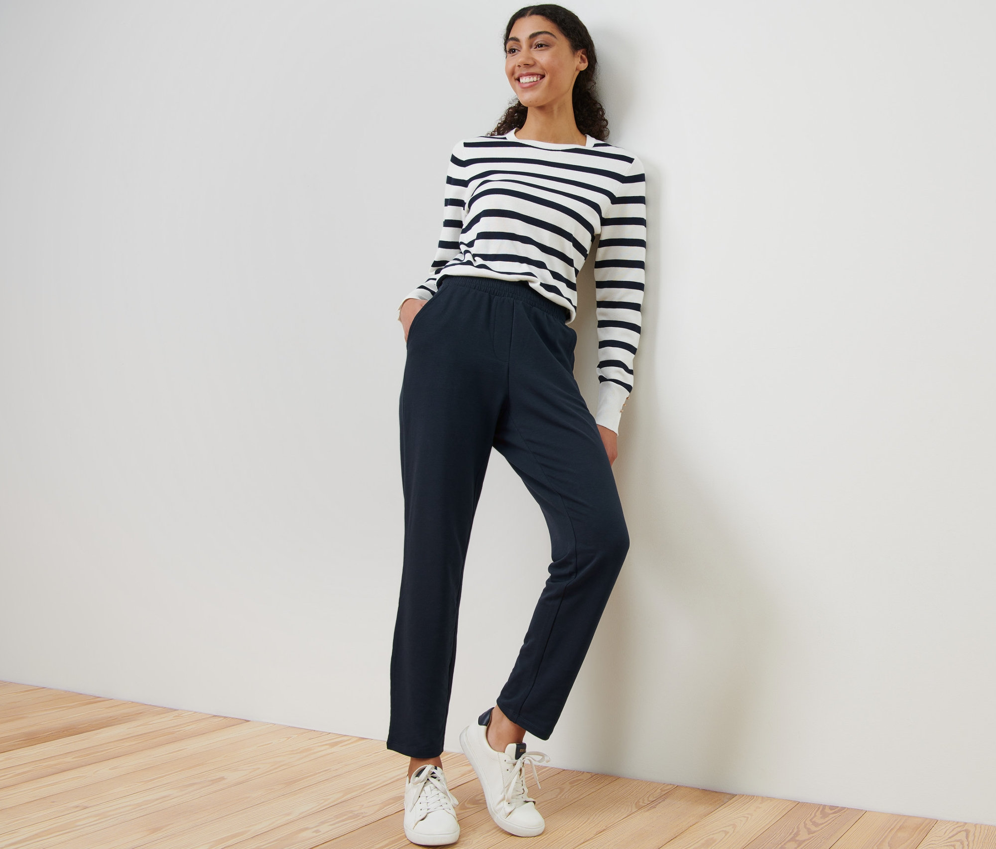 Eine Frau lächelt und lehnt an einer Wand, sie trägt ein gestreiftes Oberteil und eine Joggpants.