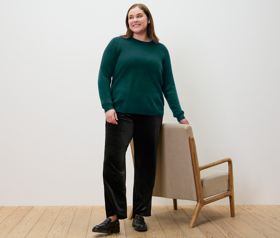 Frau steht und lehnt sich an einem Stuhl, sie trägt einen grünen Feinstrickpullover mit Cashmere.