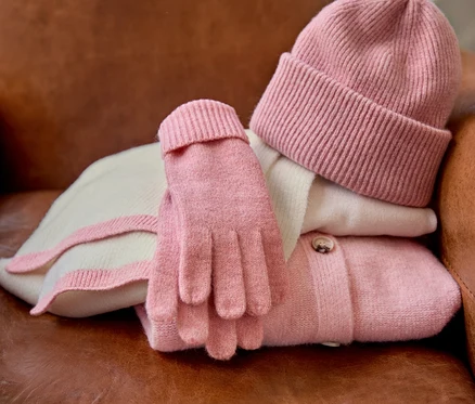 Auf einem braunen Ledersessel liegen ein Strickcardigan mit Wolle, ein rosa Feinstrickpullover mit Cashmere, eine rosé Strickmütze mit Wolle, ein Strickschal mit Wolle und rosé Strickhandschuhe mit Wolle.