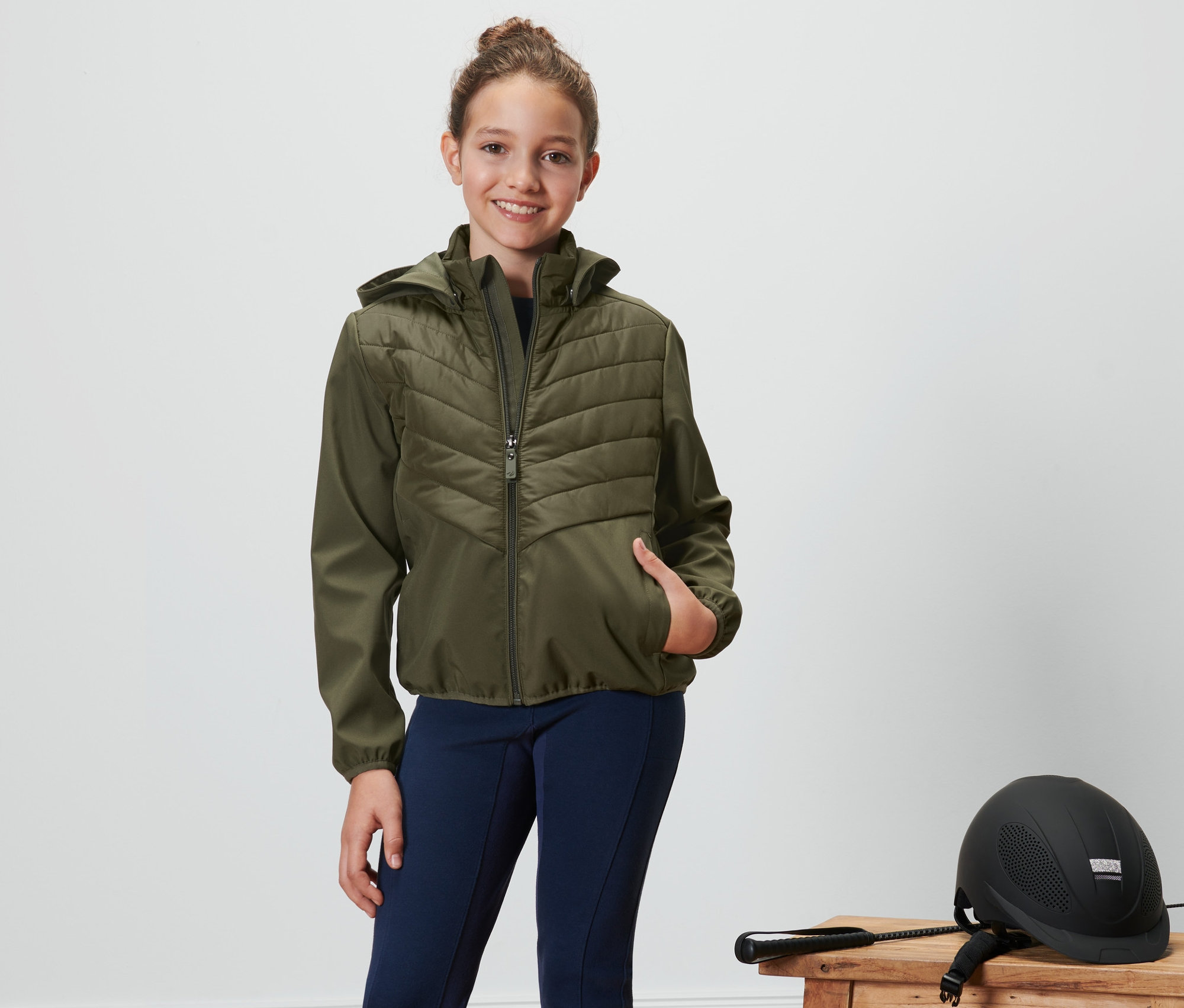 Mädchen posiert in einer Kinder-Reitjacke in Olivgrün. Daneben liegen ein Reithelm und eine Reitpeitsche auf einem Hocker.