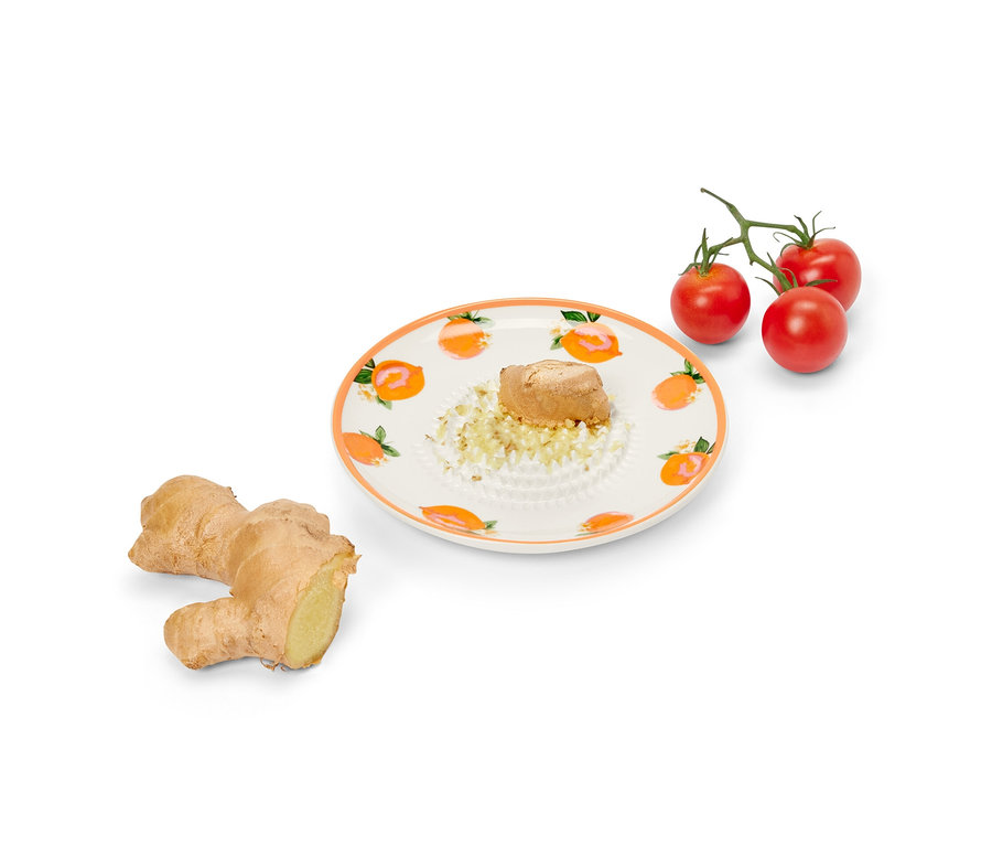 Ingwer wird auf einer Tellerreibe gerieben, daneben liegen ein ganzer Ingwer und Kirschtomaten.