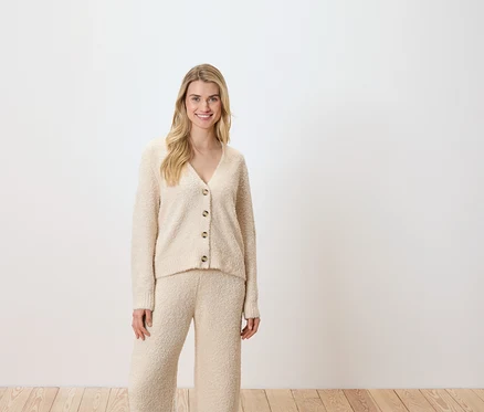 Frau posiert vor einem weißen Hintergrund und trägt eine cremefarbene Strickjacke und Hose.