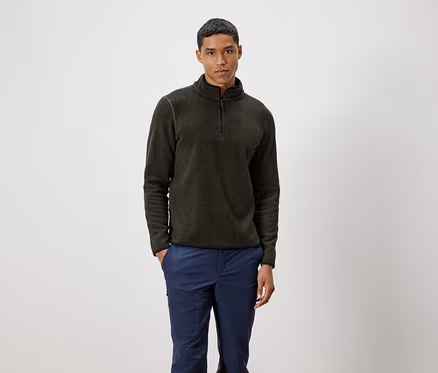 Mann steht und trägt einen olivgrünen Pullover mit Reißverschluss und blaue Hosen.