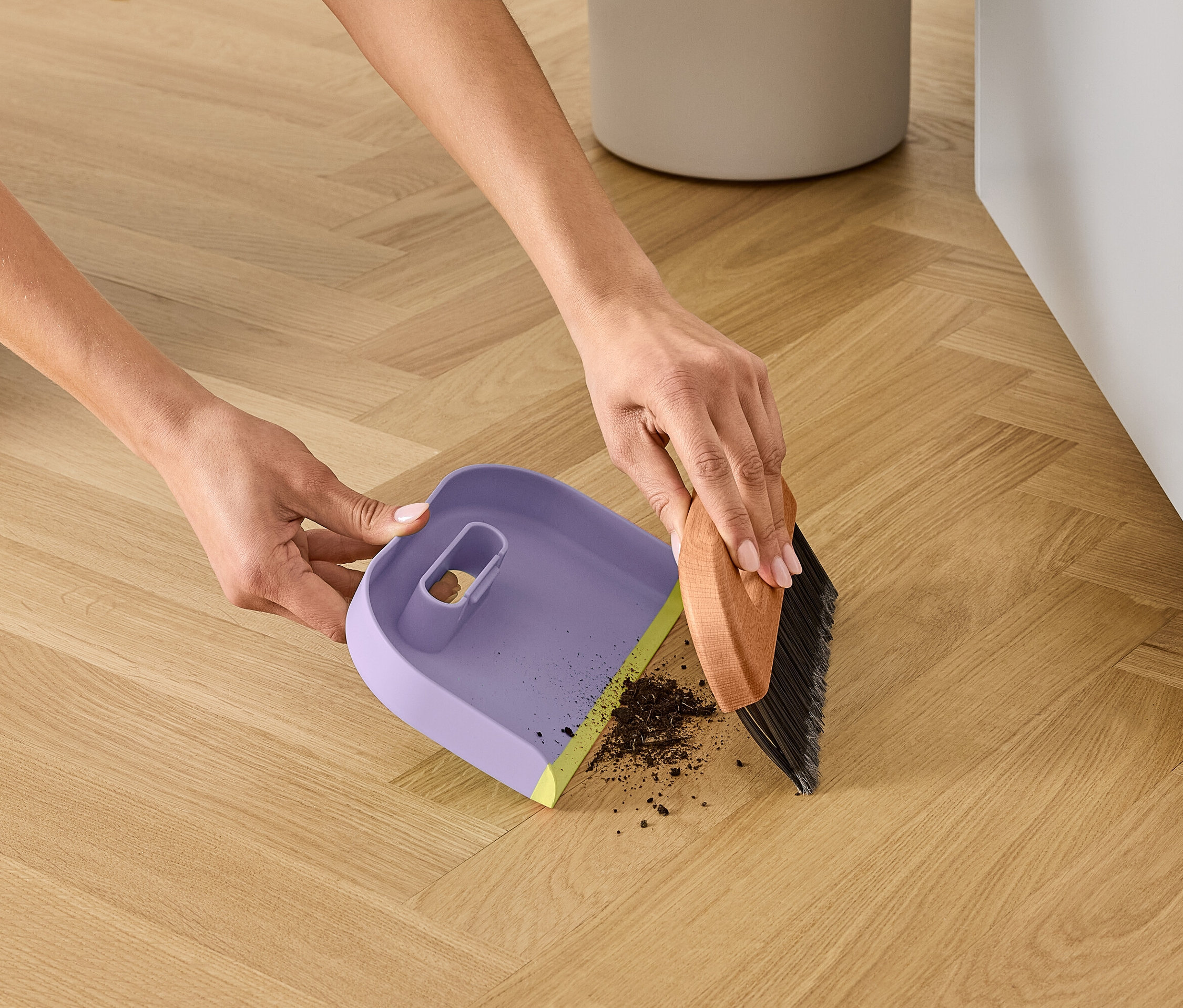 Hände kehren Schmutz mit einer braunen Bürste in eine lila Kehrschaufel mit gelbem Rand auf einem Holzboden.