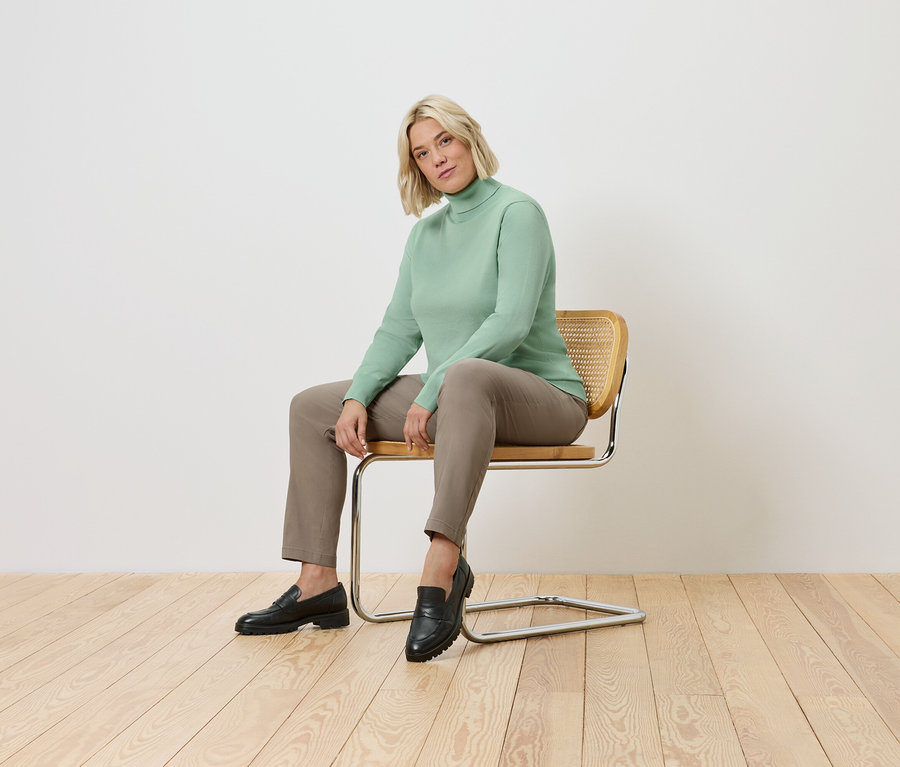 Frau sitzt auf einem Stuhl. Sie trägt einen grünen Rollkragenpullover, eine braune Hose und schwarze Schuhe.