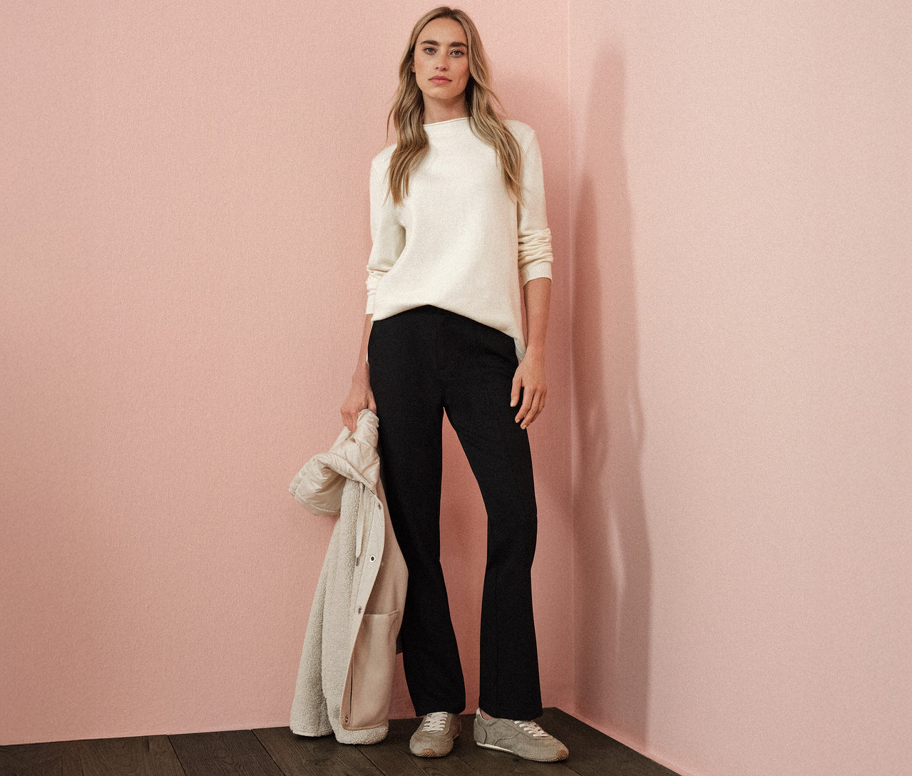 Frau trägt einen beigen Street One Strickpullover mit Rollkante, schwarze Hose und hält eine Jacke in der Hand.