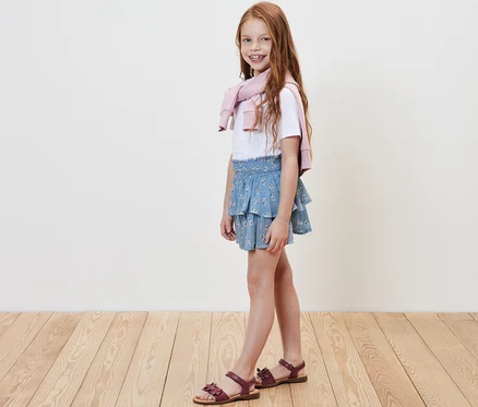 Lächelndes Mädchen posiert in Kinder-Stufenrock, weißem T-Shirt und rosa Pullover über den Schultern.
