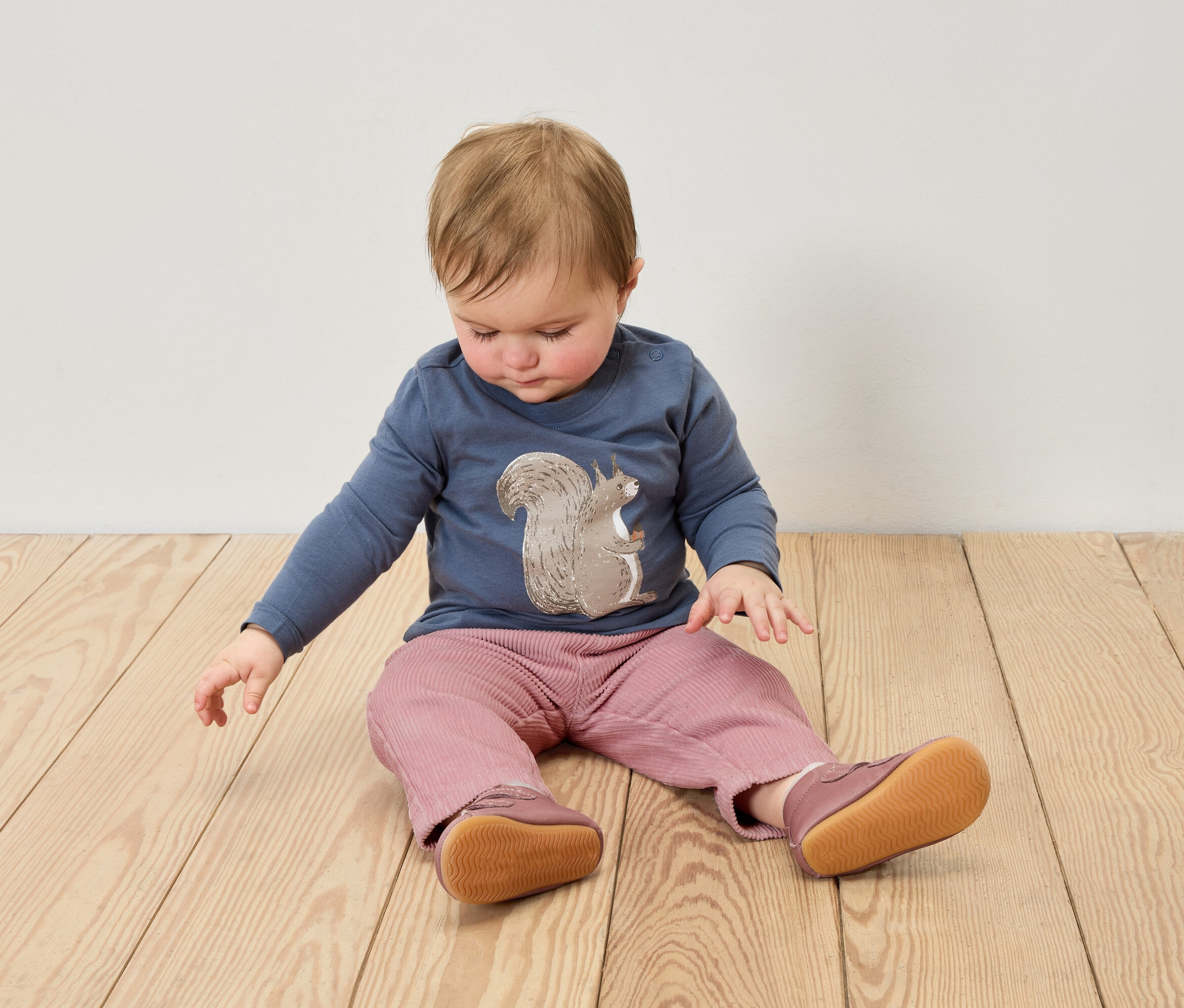 Ein Baby sitzt auf einem Holzboden und trägt ein blaues Hemd mit einem Eichhörnchenaufdruck, eine rosa Hose und Lederpuschen.