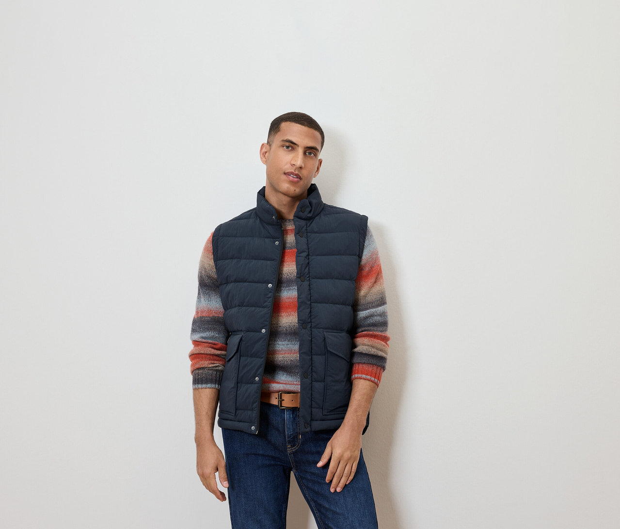 Mann trägt eine Steppweste, einen gestreiften Pullover und Jeans.