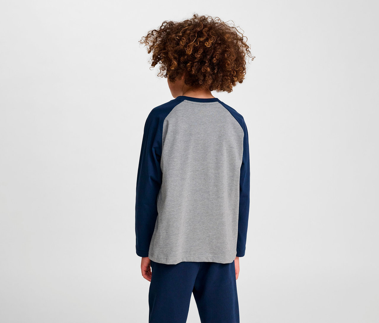 Junge mit lockigem Haar steht mit dem Rücken zur Kamera und trägt ein blaues HUMMEL HMLJR Raglan Loose T-Shirt BEE.