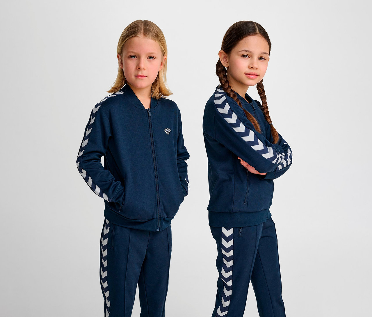 Zwei Mädchen posieren im Studio in blauen Hummel Trainingsanzügen. HUMMEL HMLJR Archive Reg Poly Zip Jacket, blau