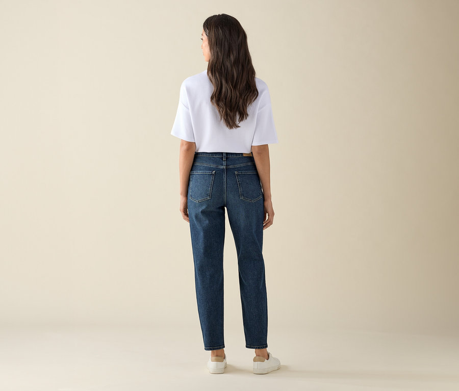 Eine Frau steht mit dem Rücken zur Kamera und trägt eine dunkelblaue Mom Jeans – Fit »Nyla« und ein weißes T-Shirt.