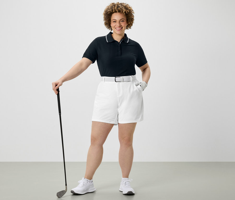 Frau hält Golfschläger, trägt dunkelblaues Funktionspoloshirt und weiße Softshell-Funktionsshorts.