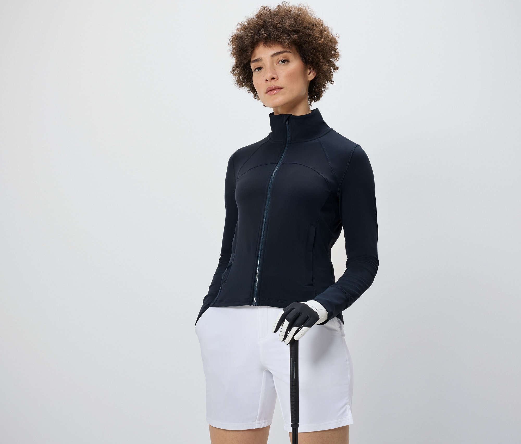 Frau trägt eine Funktionsjacke, weiße Shorts und hält einen Golfschläger.