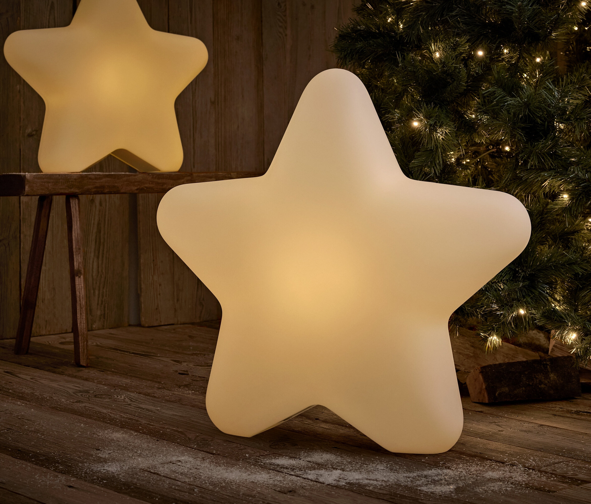Zwei sternförmige Leuchtdekorationen auf einem Holzboden vor einem Weihnachtsbaum.