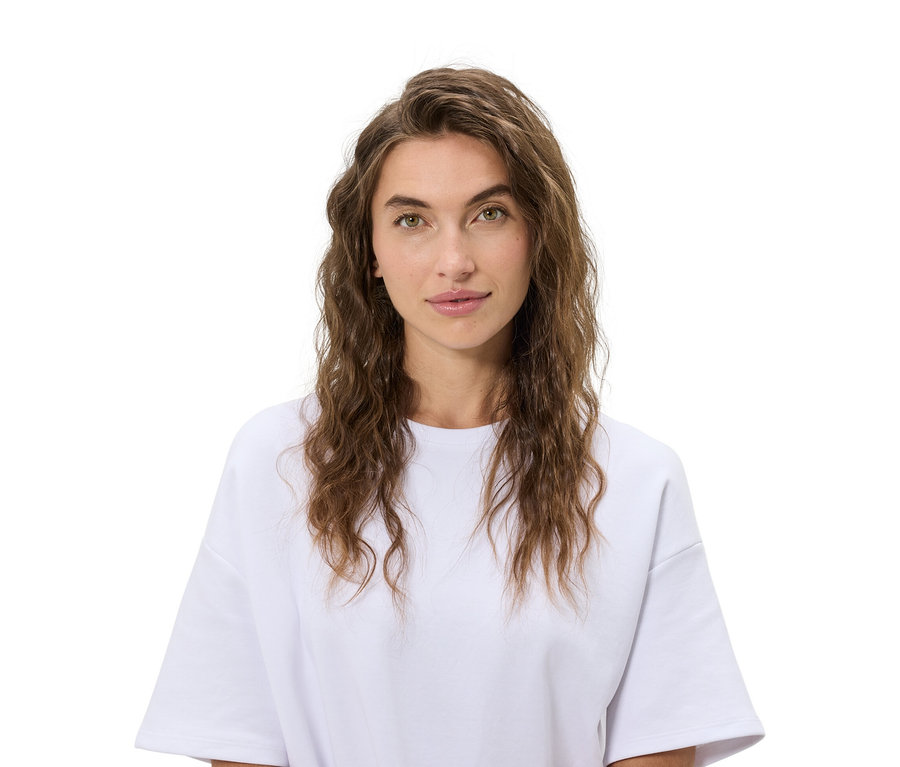 Porträt einer Frau mit gewelltem braunem Haar und einem weißen T-Shirt vor einem weißen Hintergrund.