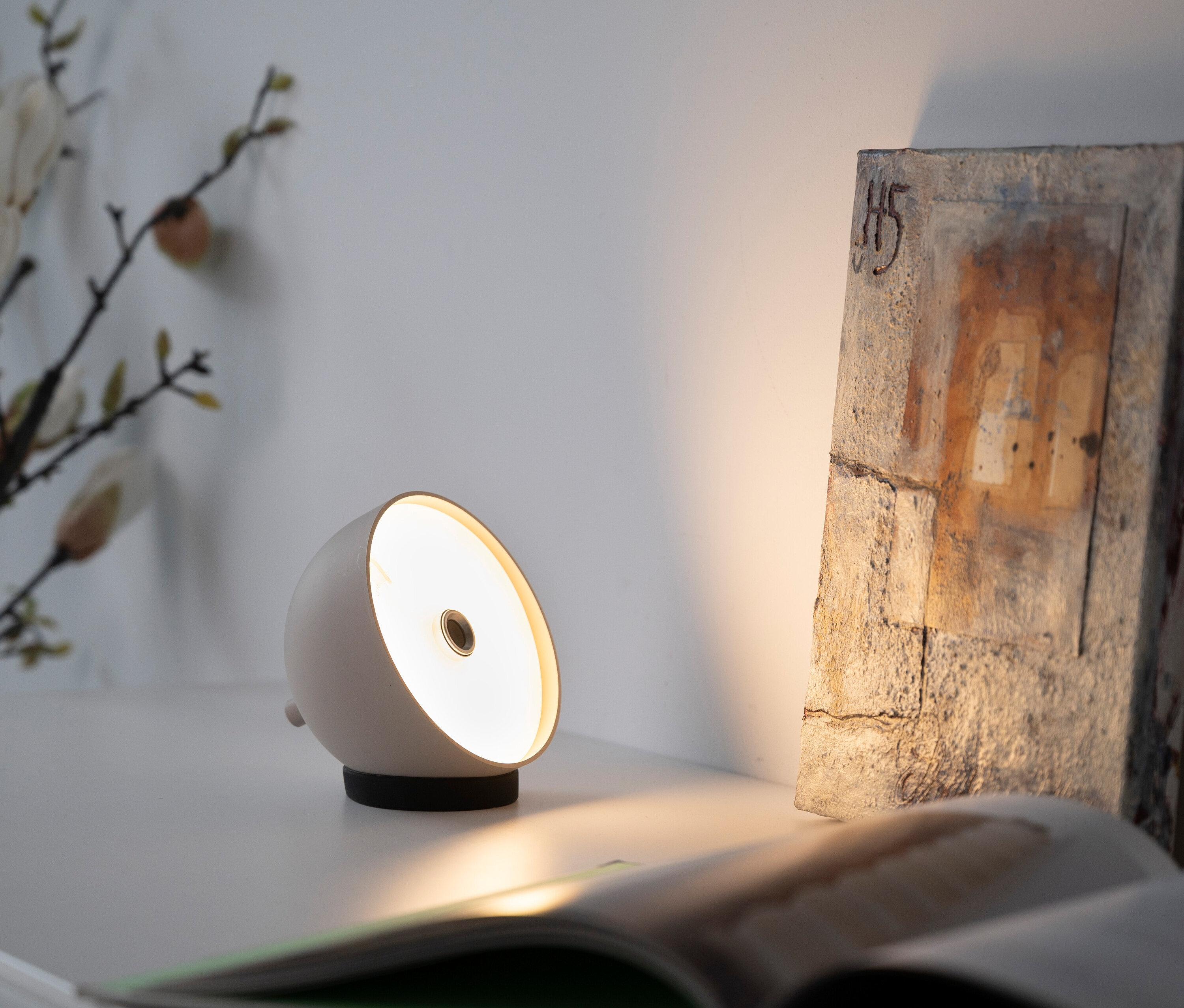 Auf einem Tisch stehen eine weiße Lampe, ein offenes Buch und ein dekorativer Stein.