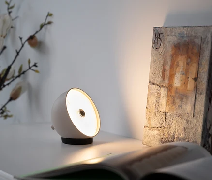 Auf einem Tisch stehen eine weiße Lampe, ein offenes Buch und ein dekorativer Stein.