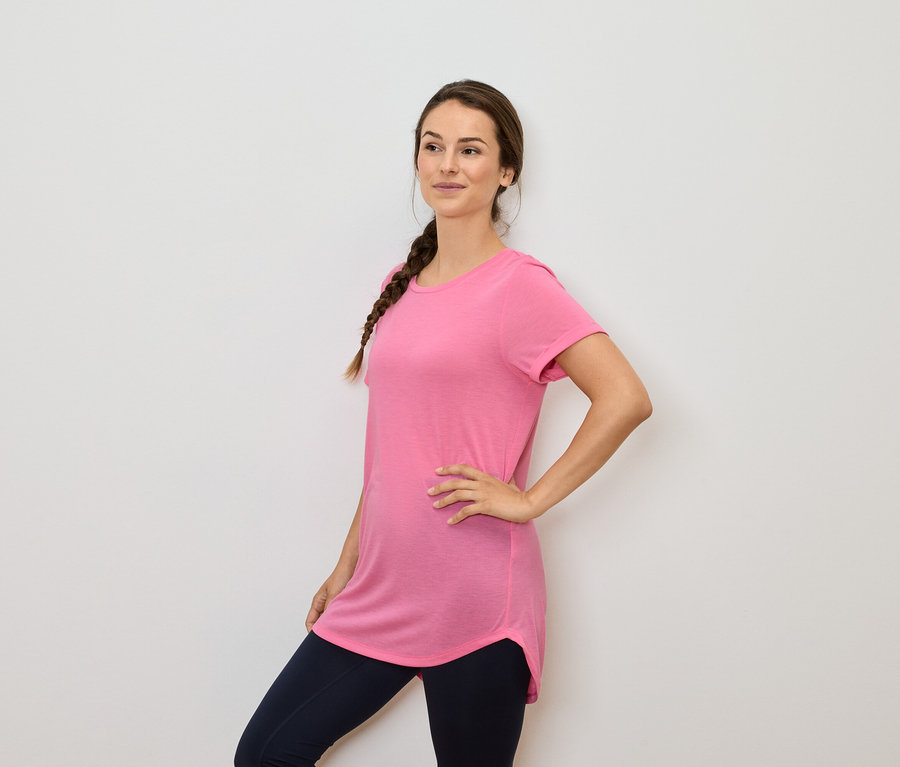 Frau mit braunen Haaren im Zopf posiert in einem rosa T-Shirt und dunkelblauen Leggings.
