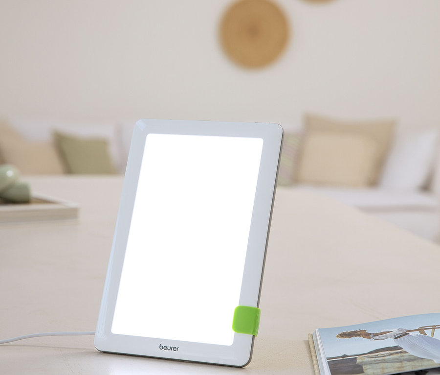 Eine weiße Lichttherapielampe mit hellem Display und grünem Clip steht auf einem Tisch.