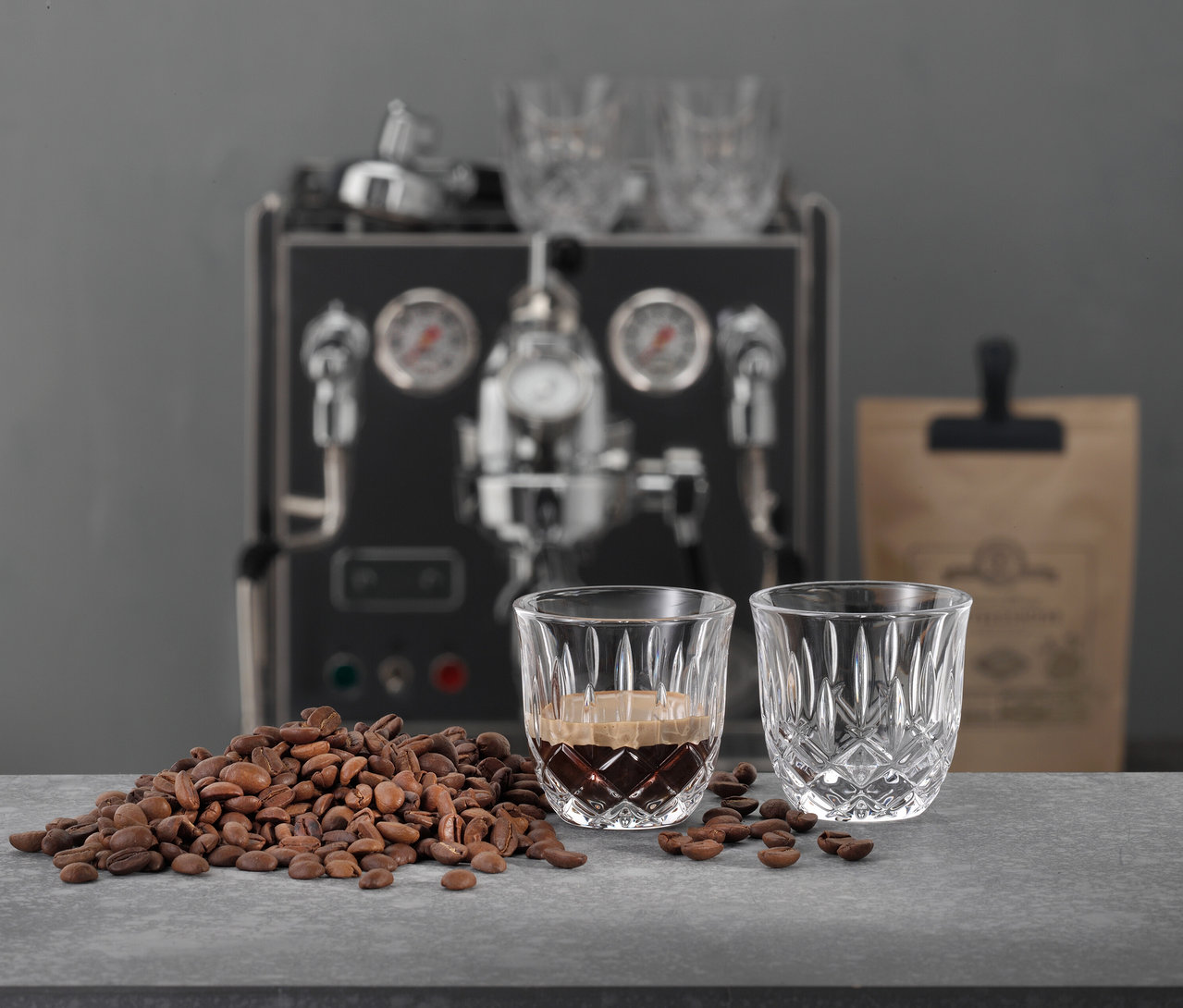 Zwei Nachtmann Espressogläser »Noblesse«, eines mit Espresso gefüllt, vor Kaffeebohnen und einer Espressomaschine.