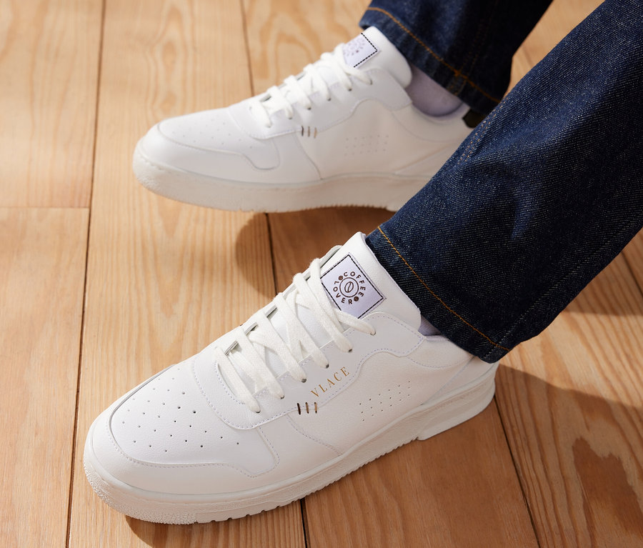 Ein Mann trägt blaue Jeans und weiße Tchibo x VLACE Coffee Lover Sneaker auf einem Holzboden.