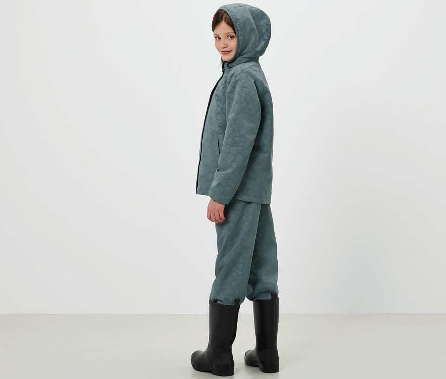 Ein Kind steht seitlich und trägt eine Thermo-Regenjacke mit Kapuze und passende Regenhose mit Gummistiefeln.