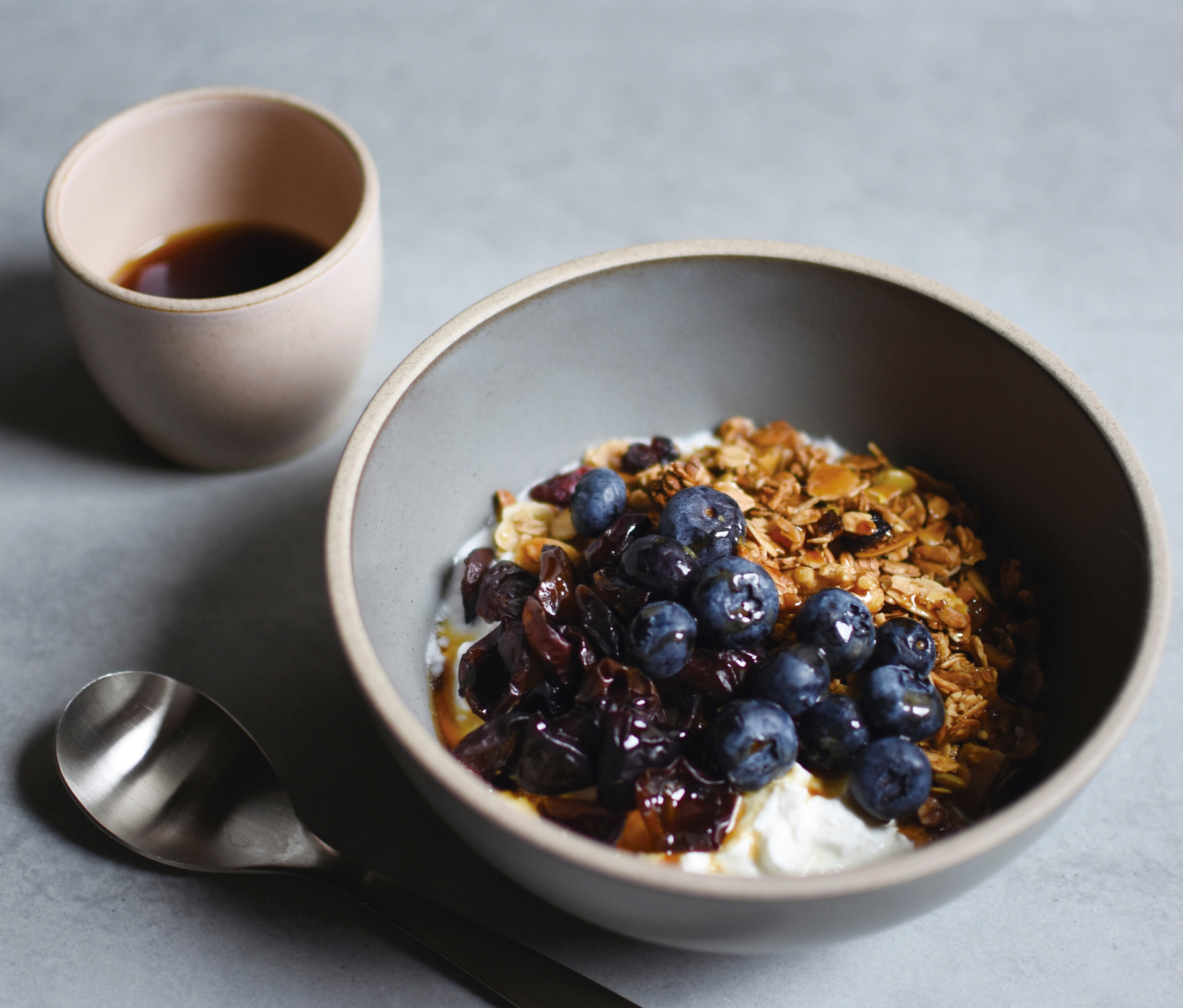 Nahaufnahme einer Müslischale mit Joghurt, Blaubeeren und Trockenfrüchten neben einer Tasse Kaffee und einem Löffel.