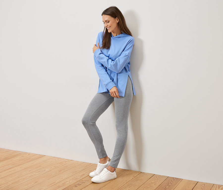 Frau lehnt an einer Wand, trägt einen hellblauen Hoodie mit Seitenschlitzen, graue Leggings und weiße Turnschuhe.