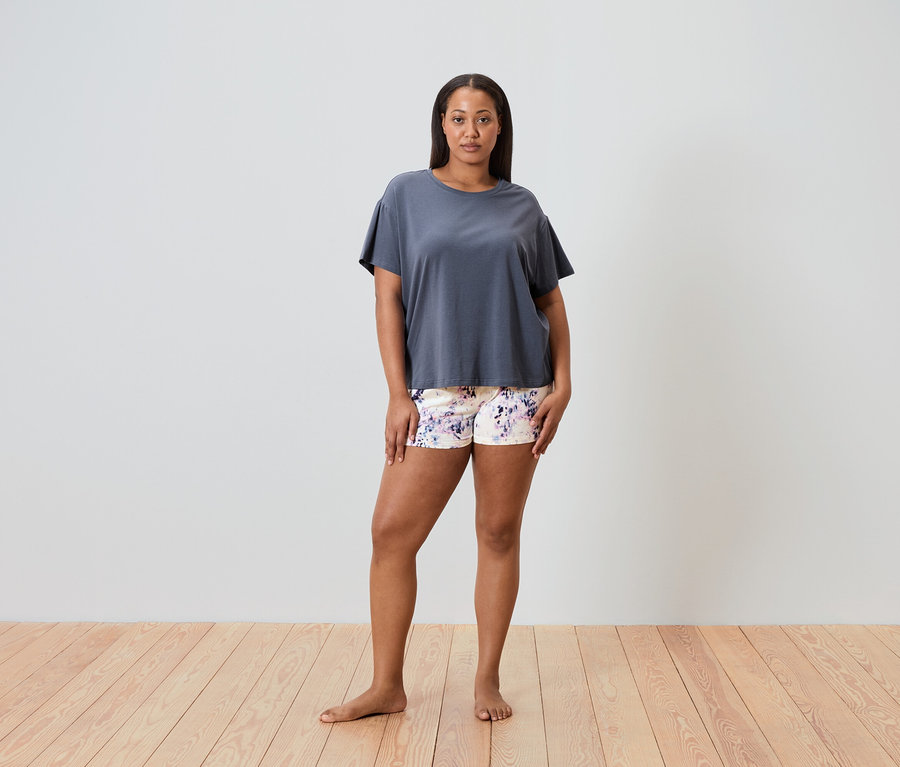 Eine Frau steht barfuß auf einem Holzboden und trägt ein graues T-Shirt und Shorts mit Blumenmuster.