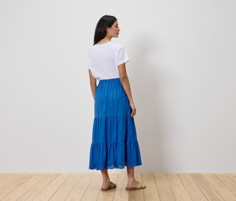 Eine Frau steht mit dem Rücken zur Kamera und trägt ein weißes T-Shirt und einen blauen, gestuften Rock.