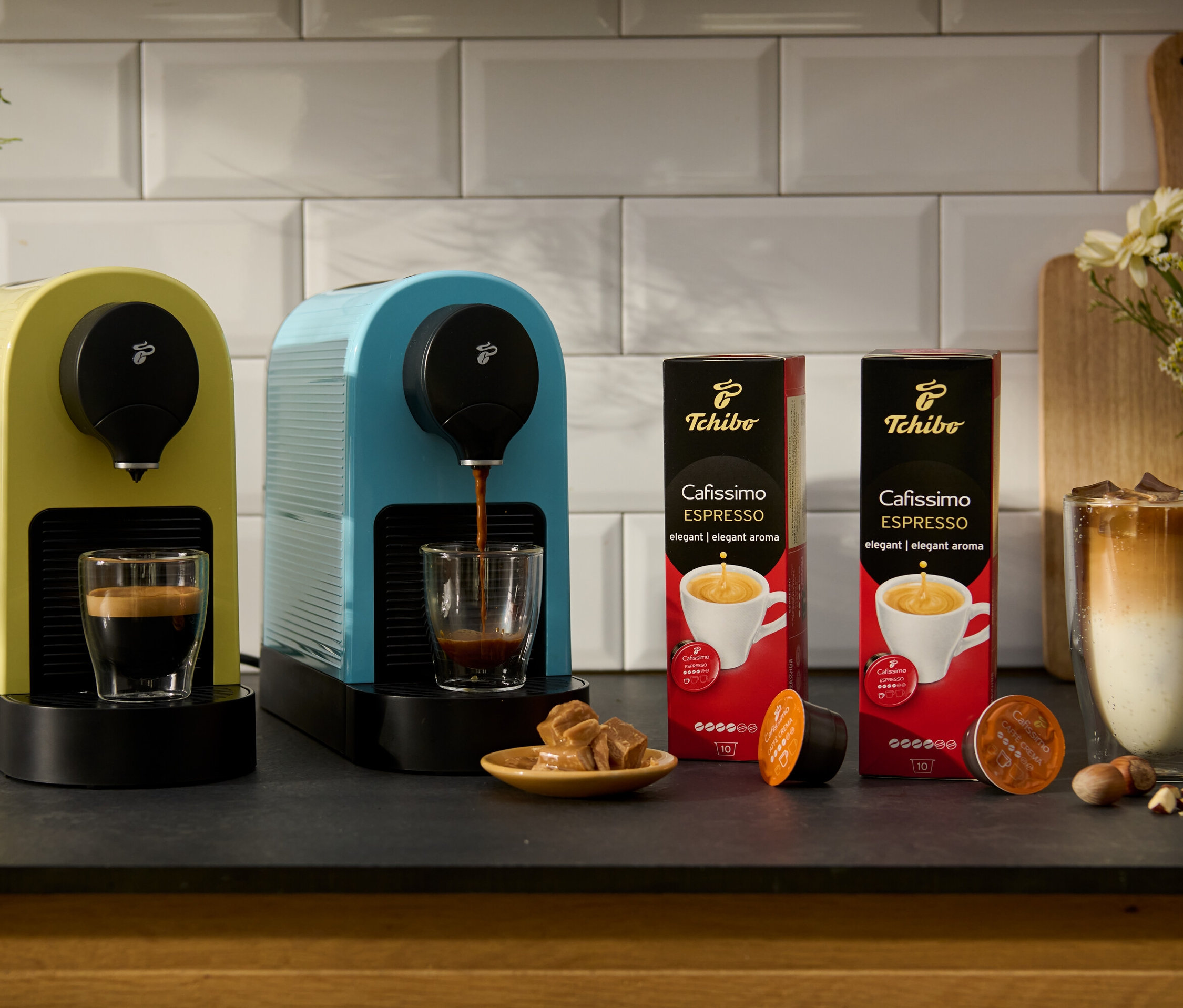 Eine gelbe und eine blaue Cafissimo pure+ Kaffeemaschine stehen neben zwei Packungen Cafissimo Flavoured Espresso - Toasted Nut.