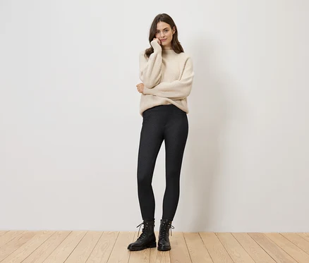 Frau in cremefarbenem Pullover, dunkler Leggings und schwarzen Stiefeln steht vor einer weißen Wand.