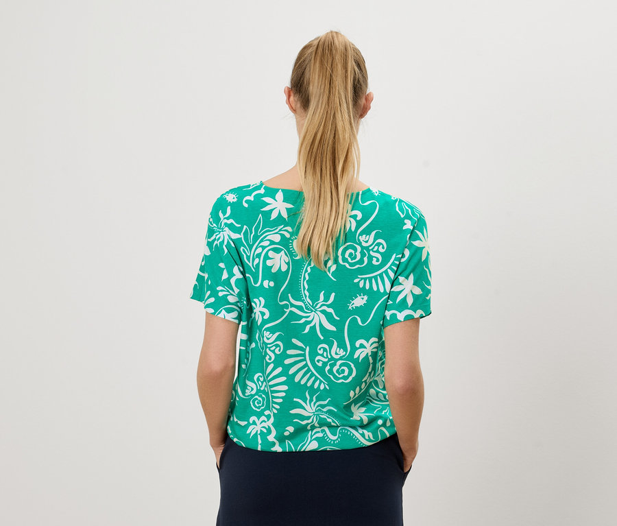 Rückansicht einer Frau mit blonden Haaren, die ein grünes T-Shirt mit weißem Muster und einen marineblauen Rock trägt.