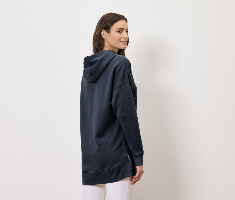 Frau trägt einen langen marineblauen Kapuzenpullover und weiße Hosen, von der Seite betrachtet.