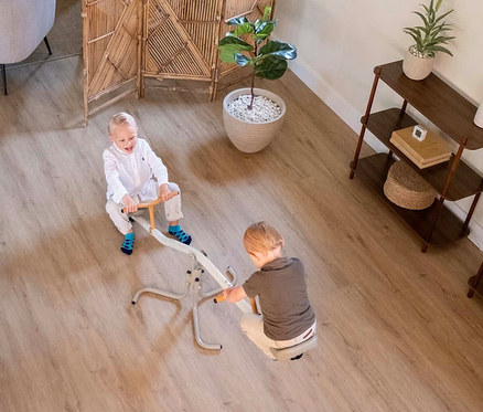 Zwei Kinder spielen auf einer Wippe in einem Wohnzimmer mit Holzboden, einem Raumteiler und Pflanzen.