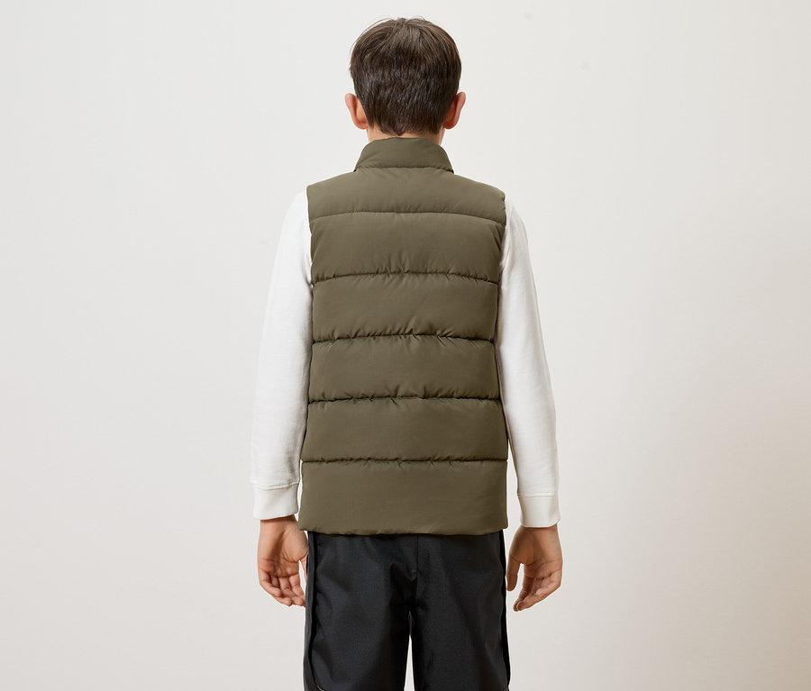 Rückansicht eines Kindes mit weißem Pullover, schwarzer Hose und olivgrüner Steppweste vor einem weißen Hintergrund.