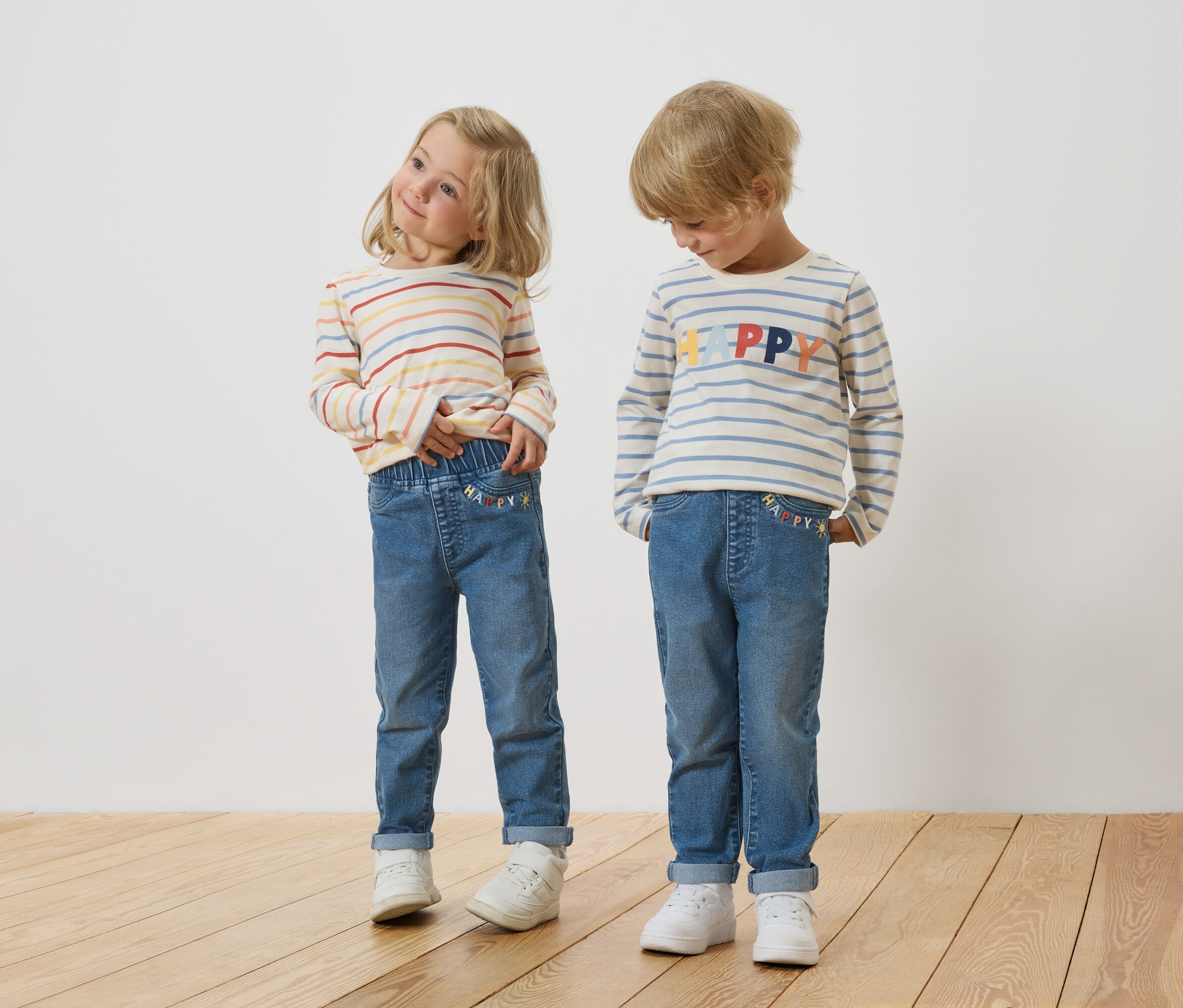 Ein blondes Mädchen und ein blonder Junge stehen nebeneinander und tragen gestreifte Langarmshirts und Jeans.