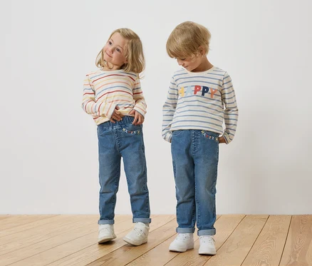 Ein blondes Mädchen und ein blonder Junge stehen nebeneinander und tragen gestreifte Langarmshirts und Jeans.