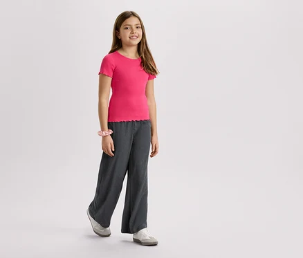 Mädchen trägt ein pinkes T-Shirt, graue Hose und Turnschuhe vor einem weißen Hintergrund.