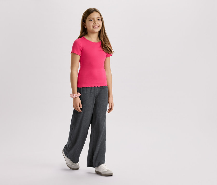 Mädchen trägt ein pinkes T-Shirt, graue Hose und Turnschuhe vor einem weißen Hintergrund.