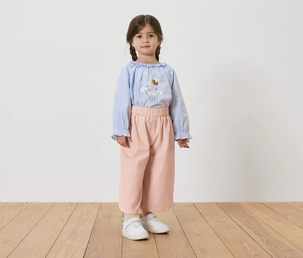 Mädchen mit Zöpfen, trägt eine hellblaue Bluse mit Blumenstickerei, rosafarbene Hose und weiße Turnschuhe.