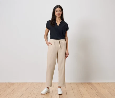 Eine Frau steht vor einer weißen Wand. Sie trägt ein schwarzes T-Shirt, eine beige Hose und weiße Turnschuhe.