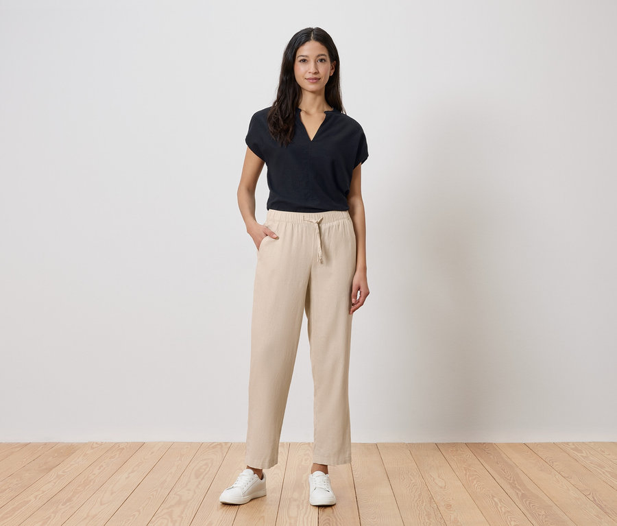 Eine Frau steht vor einer weißen Wand. Sie trägt ein schwarzes T-Shirt, eine beige Hose und weiße Turnschuhe.