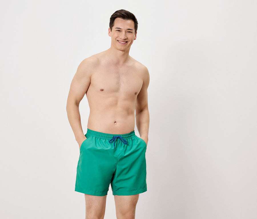 Mann mit freiem Oberkörper und grüner Badehose mit blauen Kordeln, die Hände in den Hosentaschen, vor einem weißen Hintergrund.