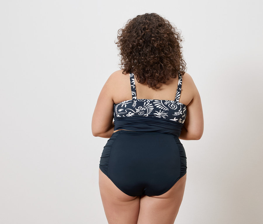 Rückansicht einer Frau mit lockigem Haar, die ein marineblaues Tankini-Top mit weißem Muster und eine marineblaue, hochtaillierte Bikinihose trägt.